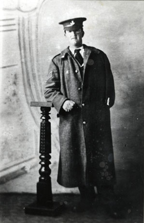 Photograph, full-length. taken in a photographer's studio of a soldier wearing a greatcoat with sergeant's stripes on the right sleeve; his left sleeve appears to be empty and tucked into his left pocket; he is wearing a cap bearing the badge of the Durham Light Infantry; he has been identified as Sergeant Joseph Cannon of Hawthorn Village