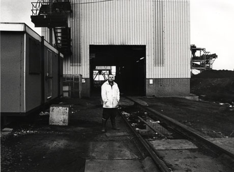 Photograph showing a man, wearing an anorak, standing beside a rail, which runs into the entrance of a large building of corrugated iron which is behind him; a fire escape can be seen on the exterior of the large building behind a small temporary building, which is at the left of the photograph; a metal structure can be seen at the right of the picture; the buildings are industrial, and may be colliery buildings, at Hawthorn