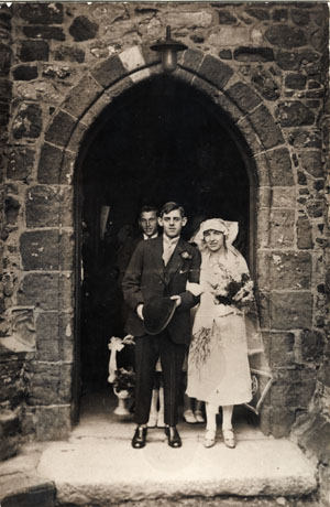 Photograph of a bride and groom at the entrance to Monk Hesledon parish church after their wedding; the photograph shows the arch of the church, the bride in her gown, carrying a bouquet and the groom in a suit, carrying his hat; behind the groom the face of a young man can be seen and the shoes and legs of the bridesmaids