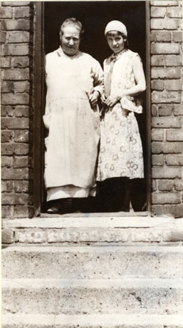 Photograph showing a middle-aged woman wearing a voluminous pinafore standing in the doorway of a house accompanied by a young woman wearing a sleeveless patterned dress and light-coloured hat; the photograph is taken close-up and shows the women, the doorway and two of the steps leading to the door; the photograph has been identified as depicting a scene in Hesledon