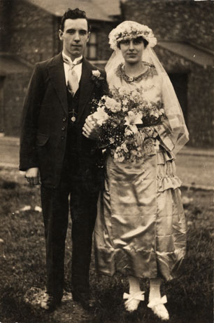 Photograph of a bride and groom taken in the open air with a house in the distance behind them; the bride is wearing a satin dress and carrying a bouquet of lilies; the hem of her dress about six inches above the ground revealing large bows on her shoes; her headdress is in the style of the 1920s; the groom is wearing a dark suit with a wing collar; the photograph is described as Hesleden Church Wedding
