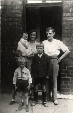 Photograph showing a man and a woman in workaday clothes standing in front of the doorway of a brick house, with three small boys of the ages of approximately four, six and ten years; the woman is holding an infant of a few months; the photograph is described as Family, Hesleden, 1940s