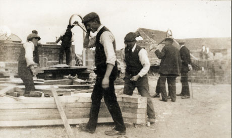 Photograph of seven men wearing waistcoats and caps, working on wood lying on the ground in the foreground, and on walls in the background; men can also be seen on a building in the background at the right of the picture, which has been identified as Building The Ship Inn, Hesledon