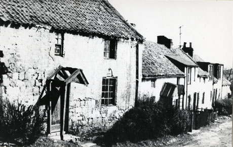 Photograph of the exterior of cottages at Monk Hesleden in a dilapidated state; the photograph shows a row of cottages and the cottage closest to the camera is a building of rough stone covered in rough render, with three small windows and a broken porch over the door in the middle of the building; the road in front of the cottages appears to be un-made-up; the photograph was taken immediately before the cottages were demolished; the photograph is described as Monk Hesleden Cottages Before The Bulldozers