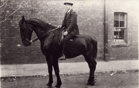 Photograph of a young boy, in suit and flat cap, sitting on the back of a horse of dark colour; behind the horse and boy the wall of a brick house can be seen; the photograph is described as Tot Carling, 1911