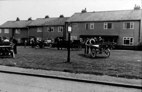 Photograph of an open space of grass with three semi-detached houses behind it; on the green are horses harnessed to four carts; the photograph has been identified as The Village Green, Sunday Morning, High Hesledon