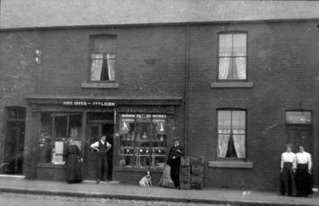 Photograph showing the windows of a shop with a man and a woman standing in the doorway; above the doorway are the words Post Office Hesledon; in the right hand window are advertisements for Rowntree's Cordial; a man and a dog are standing at the right of the right-hand window; two women, wearing dark skirts and light blouses, are standing in front of the doorway of the adjoining terraced house