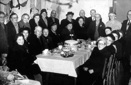 Photograph showing eleven elderly men and women sitting round a table laid with cups and saucers, food and an elaborate celebration cake; behind them twelve other men and women are standing; all of them are wearing coats and hats; a streamer can be seen at the extreme left of the picture; the picture has been identified as Over 60s Party, Hesledon