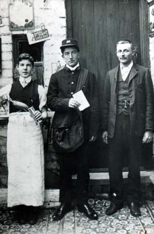 Photograph showing two men and a boy standing together in a row, inside a building, with a light-coloured wall and a wooden door behind them; they are standing on a patterned floor; on the left is a boy wearing a waistcoat and a long apron, holding a scoop putting something in a paper container held in his left hand; in the centre is a man dressed in a dark uniform with a bag over his shoulder; at the right is a man dressed in a suit and tie; the photograph has been identified as Mr. Scott, Postmaster, Hesledon