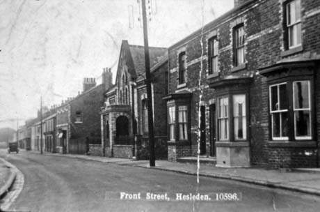 Postcard photograph entitled Front Street, Hesledon. 10596, showing one side of the street; nearest the camera are three terraced houses with bay windows; next to them is a chapel followed by blocks of terraced houses with one or two shops in them; a car can be seen in the distance driving away from the camera