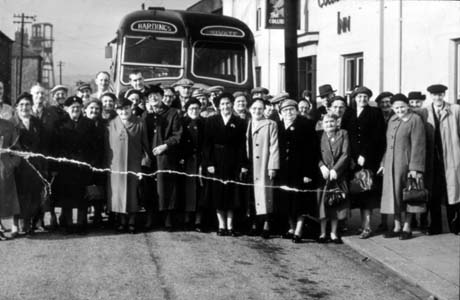 Photograph showing approximately forty elderly men and women standing in a group before the front of a single-decker bus, which has Hardings and Private on its indicator boards; the people are all wearing coats and hats; behind them and the bus, the winding gear of a colliery can be seen; the man at the extreme right of the group, wearing a mackintosh and a cap, may be the driver of the bus; the bus is parked beside the Colliery Inn; the photograph has been described as Over 60s Outing, Hesledon
