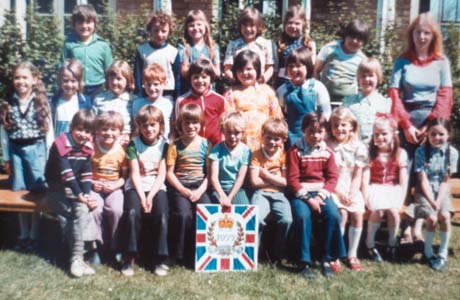 Photograph of twenty four children, aged between approximately eight and nine years, posed in three rows outside a building; a woman is standing with them; they have a board in front of them with a Union Jack on it on which has been superimposed a crown, laurel wreath and the figures, 1977, to commemorate the Silver Jubilee of Queen Elizabeth II; the children have been identified as Classes 1 and 2 of a Junior School in Hesleden