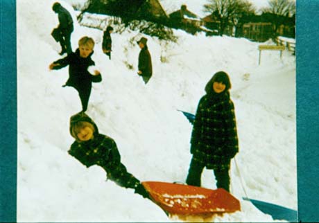 Photograph of two girls, aged approximately twelve and eight years, with a toboggan, on a slope covered in snow; behind them are four other children on the slope and semi-detached houses in the distance; they have been identified as Emma and Vickie Hunt in Hesledon in 1979, when the village was cut off by snow