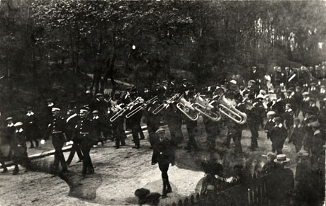 Photograph showing, rather indistinctly, members of a band in uniform walking along a road with tall trees in the background; the front rows of the band are playing brass instruments and are preceded by two men in uniform; the bandsmen are followed by a parade of men in the same uniform and are being watched by spectators on either side of the road; the photograph has been identified as Band Leading Procession, Blackhill's Terrace, Hordern, 1913