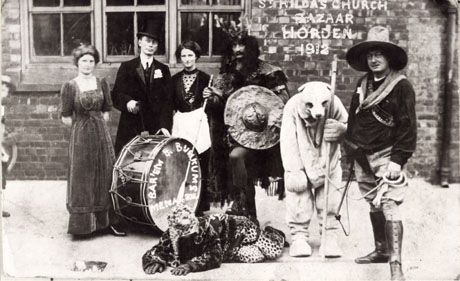 Photograph seven men and women in fancy dress posed in front of a building on which has been written the words St. Hilda's Church Bazaar, Horden, 1912; the people are dressed as follows: one woman in a formal evening dress; a man in a suit and bowler hat; a man dressed as a warrior with a shield; an individual dressed as a bear; a man dressed as an American showman; an individual dressed as a leopard; also to be seen in the picture is a large drum on which is written: Bar'em & Bunkums Menagerie