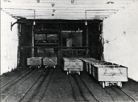 Photograph showing three rows of trucks in a tunnel on rails, drawn up before a barrier behind which more trucks may be seen; the walls and ceiling of the tunnel are painted a light colour and two lights are suspended from the ceiling; the photograph hads been identified as Shaft Bottom, Hutton Seam, Horden
