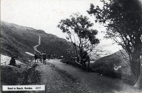 Postcard photograph entitled Road to Beach, Horden. 4227, showing the surface of a track in the foreground and it winding away into the distance over a hill to the left of the picture; to the right, a vista of countryside may be seen; a number of boys, who may be boy scouts, are walking along the road away from the camera; two boys are sitting either side of the road in the foreground