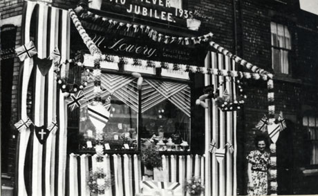 Photograph of the facade of the shop belonging to M. Lowery, Fruiterer and General Dealer, Horden, decorated to mark the Silver Jubilee of King George V; above the window of the shop is written: 1910 Silver Jubilee 1935 and the window is elaborately decorated with flags, shields, balloons, hanging baskets, crowns, flowers, ribbons and streamers; a woman in a flowered dress is standing in front of the shop