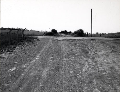 Photograph of the surface of a road leading from the camera into the distance; a road can be seen crossing the road from right to left of the picture; in the far distance the roofs of post Second World War houses can be seen; the photograph has been identified as Road leading from Warren House to Horden Hall Farm. (Crossed by Coast Road 1976)