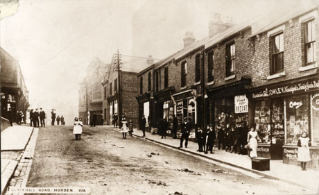 Postcard photograph entitled Blackhill Road, Horden. 1566, showing the right-hand side of a road going uphill away from the camera; the facades of shops can be seen, including those of R( )owley, Tobacconist, Newsagent, Fancy Goods Dealer, and Gales Penny Bazaar; in the distance is a large building, possibly a cinema; approximately twenty six indistinct figures can be seen in the road and on the pavements