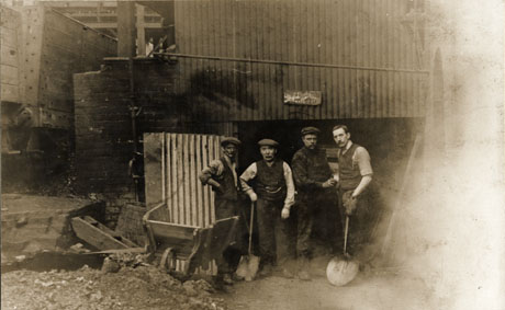 Photograph of four men standing at the entrance of a building at a colliery, identified as Horden; all are wearing working clothes and two are holding shovels; the building appears to be constructed of corrugated iron; a large truck can be seen on the left of the picture and a pair of small steps, a barrel and a wheelbarrow are lying near the men, who have been described as Coke Oven Officials
