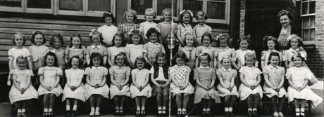 Photograph of thirty four small girls of approximately six years of age, posed outside a brick and wood building with a woman, presumably their teacher; the photograph is described as Horden Junior Girls' School, 1955