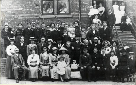 Photograph of fifty people and ten children grouped outside a terraced brick house with high steps leading to the entrance; people can be seen in the windows of the house; two soldiers can be seen on the front row of the group and what appears to be the cake can be seen on a table in the middle of the front row; all the people are dressed formally; the occasion has been described as Mrs. Gilchrist's Wedding, Blackhills Terrace,Horden, 21 September 1914