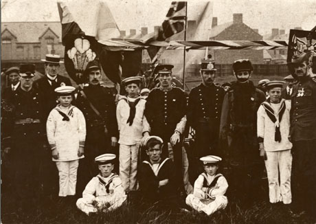 Photograph of eight men and five boys dressed as members of the armed forces of different countries; behind the men and boys, the rear of a horse and part of a float with Union Jacks and the Prince of Wales feathers can be seen; four other people and the facade of a large building, possibly a school, and the end of a terrace of houses can also be seen; the photograph has been identified as Carnival Float Advertising The Forces, Horden, 1920s
