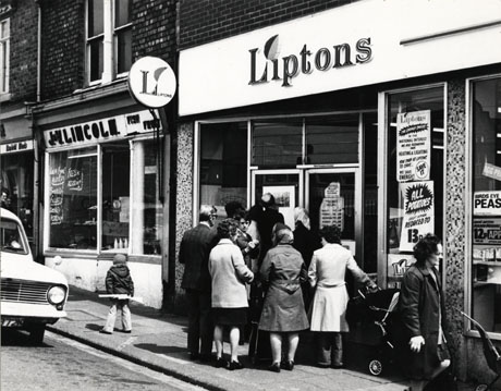 Photograph showing, close up, the facade of a Liptons shop in Horden; only the facades of Liptons and of the neighbouring fishmonger, J. and V. Lincoln, can be seen; outside Liptons a group of eight people with their backs to the camera are waiting for the shop to open; according to a notice on the door, the shop opened at 9.45 in the morning; also according to a notice in the window of the shop its proprietors were reducing the heating and lighting in the shop in the National Interest; the date of the photograph has been given as 1976; the front of a car can be seen to the left of the photograph and a woman can be seen walking on the right of the photograph; a child with a toy gun is standing with his back to the camera in front of the fishmonger's shop