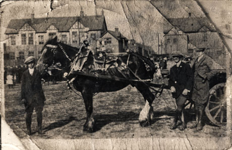 Photograph of a horse between the shafts of a cart, which is out of shot, with two men leaning against the cart and one man holding the horse's head; behind the horse is, in the distance, a large half-timbered house and a street of houses can be seen receding into the distance; indistinct figures can be seen behind the horse and men; the photograph has been identified as Carnival Horse, Outside Horden Club