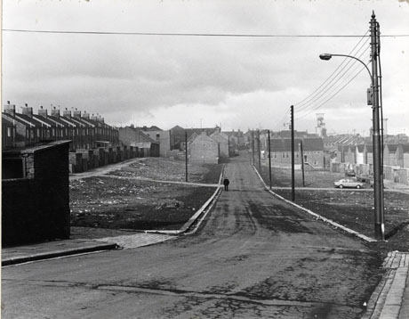 Photograph showing a road receding from the camera, with a row of terraced houses on the left and the backs of houses on the right; immediately on either side of the road are patches of bare earth where houses identified as 4th Street, Horden, have been demolished; a car can be seen in the distance on the right and above the roofs of the houses on the right the winding gear of the colliery