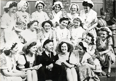 Photograph of seventeen members of the Horden Operatic Society dressed as milkmaids surrounding a man dressed as a naval officer; in the distance, on the right, two men dressed in oriental costume, may be seen; the group is posed outside a brick building; the photograph is dated 9 September 1946