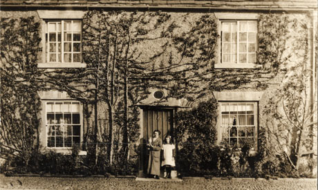 Photograph of the facade of a building with four sash windows and a doorway with stone lintel with a round fanlight; the building is covered with creeper and there are a woman, wearing an ankle-length coat and carrying a cat, and a girl, aged approximately twelve years, wearing a pinafore and boots, standing in the doorway; the building has been identified as Ellisons Farm, Horden