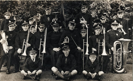 Photograph of seventeen men and boys, members of the band of the Horden Branch of the Salvation Army, posed against trees; fourteen are holding instruments; one is holding a conductor's baton; one is holding a banner; and one appears to be the leader of the group; two boys sitting on the ground have no instruments and one is holding a triangle