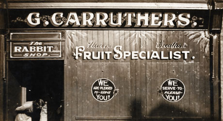 Photograph of the facade of the shop belonging to G. Carruthers, with the following notices on the facade:  The Rabbit Shop Flowers Wreaths &c. Fruit Specialist We Are Pleased To Serve You; a woman can be seen bending over in the doorway