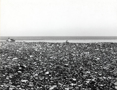 Photograph of the beach at Horden with the sea in the distance and with a man sitting in the distance near the shoreline with his back to the camera, and a motor scooter parked near the shoreline; the beach appears to consist of stones or shingle