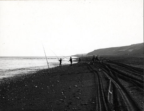 Photograph looking along the beach at Horden showing the sea to the left of the picture and hills to the right of the picture; in the centre of the picture, the beach with tyre marks along it and stones lying on it can be seen; in the middle distance, seven men can be seen fishing in the sea; they have been identified as members of Horden Angling Club