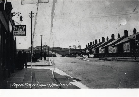 Photograph entitled Aged Miners' Homes Horden 43, showing a road descending a hill towards the camera with aged miners' homes on its right- hand side at the lower part of the hill; beyond the Homes, open fields can be seen; on the left-hand side of the road further low buildings, possibly Aged Miners' Homes, can be seen half way up the hill, and a hanging sign advertising Wills's Capstan Navy Cut Cigarettes can be seen on a building at the bottom of the hill' which cannot be identified; two indistinct men can be seen walking away from the camera on the left-hand side of the road