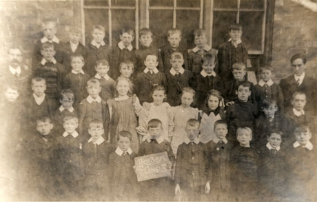 Photograph of thirty eight children, aged approximately eight years, posed against a brick wall and window, accompanied by two men, presumably the childrens' teachers; a child on the front row is holding a notice reading Horden Group II