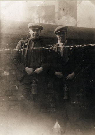 Photograph of two men standing in front of a wall wearing caps, scarves, jackets, and trousers, and carrying miners' lamps; both have a badge pinned to their lapels; they have been identified as Ralph and Bill Porter of Horden
