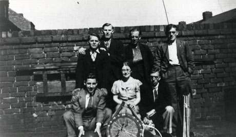 Photograph of six men and one woman posed in front of a brick wall; un front of the woman are a tennis racquet, a darts board, and another unidentifiable racquet; in front of one of the men is a large ball; one man has his arm in a sling; the photograph has been identified as 7th Street, Horden