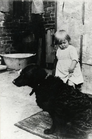 Photograph of a child, aged approximately three years, standing behind a large dog with a curly coat, which is sitting on a mat; behind the child and dog a small tin bath and the corner of two brick walls can be seen; the photograph has been identified as Backyard, 7th Street, Horden