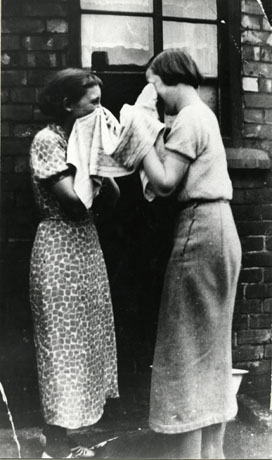 Photograph of two young women, each with the end of a small towel held to her face, standing outside a window in a brick house; one woman is wearing a skirt and short-sleeved blouse and the other a patterned dress; the photograph has been identified as Backyard Wash, 7th Street, Horden