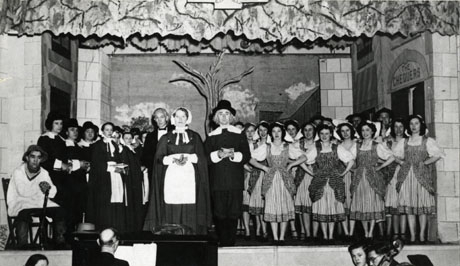 Photograph of the cast of a production of The Quaker Girl standing on the stage of the Drill Hall, Horden; the cast is in costume and the scenery of the production can be seen behind the cast