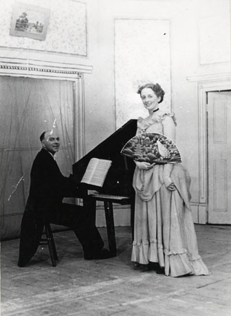 Photograph of a man in tails sitting at a grand piano, accompanied by a woman wearing an evening dress and carrying a fan; behind the couple scenery representing a drawing room can be seen; the photograph has been identified as a production of Bittersweet by Noel Coward, performed by the Horden and District Operatic Society