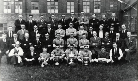 Photograph of the eleven players of the Horden Colliery Welfare Football Team posed with thirty other men in front of a large building with large windows; in front of the players are four trophies and a football