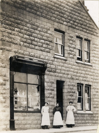 Photograph of the exterior of a stone building with five normal windows and a shop window; three women can be seen standing outside the building near the shop window; all three are wearing long skirts and aprons; the photograph has been identified as Mrs. Miller and Daughters, Miller's Cake Shop, Cotsford Lane, Horden