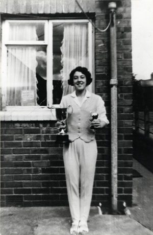 Photograph of a woman standing in front of the wall of a house with a window immediately behind her; she is dressed in trousers and waistcoat and is holding two trophies; the photograph is described as Skidmore Horden 100-200 Yards Champion