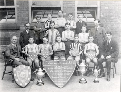 Photograph of eleven members of the Horden Council School Athletic Football Club accompanied by two other boys and four men, posed in front of a brick building; in front of the group are four trophies on the ground and four boys on the front row are holding shields; in the centre of the picture is a notice reading: Horden Council School A. F. C. 1923- 1924 Trophies: East Durham League Cup; County B.& O. Shield; Bethune Cup; Shotton Charity Cup