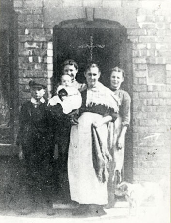 Photograph of a woman, wearing an apron and shawl, carrying a baby, accompanied by two women behind her and a boy, of approximately twelve years and wearing an Eton collar and a cap, to her right; they are posed in front of the entrance to a brick house; they have been identified as Elizabeth Byers and Family, 22 First Street, Horden